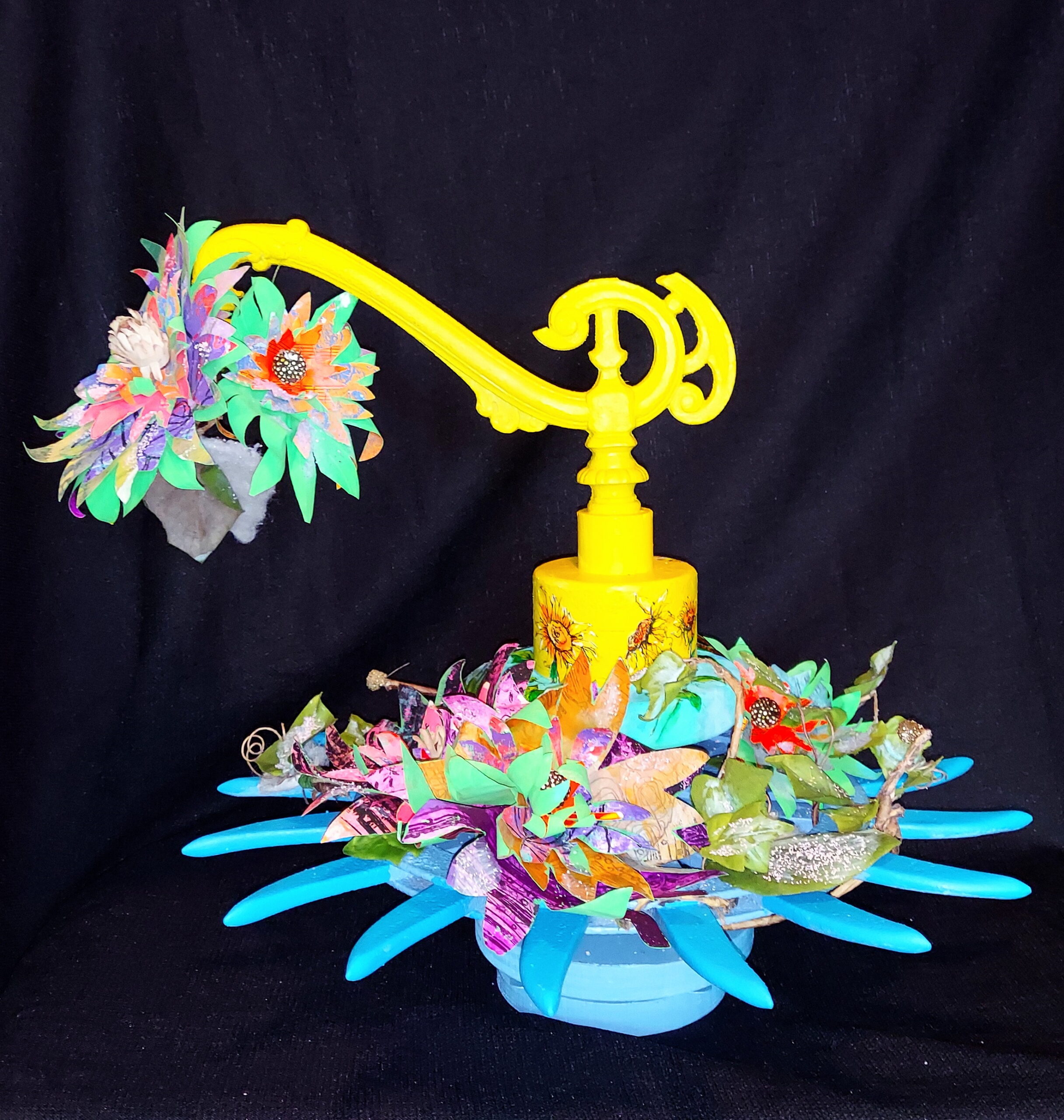 This is a photo of a sculpture with paper flowers that wrap a blue wheel that spins. The flowers are covered with a light dusting of snow which appears to be melting. This sculpture spins on its base and has a wind-up music box that plays the song Edelweiss. This piece shows how liminal art starts conversation. It raises the question, "Why?"
