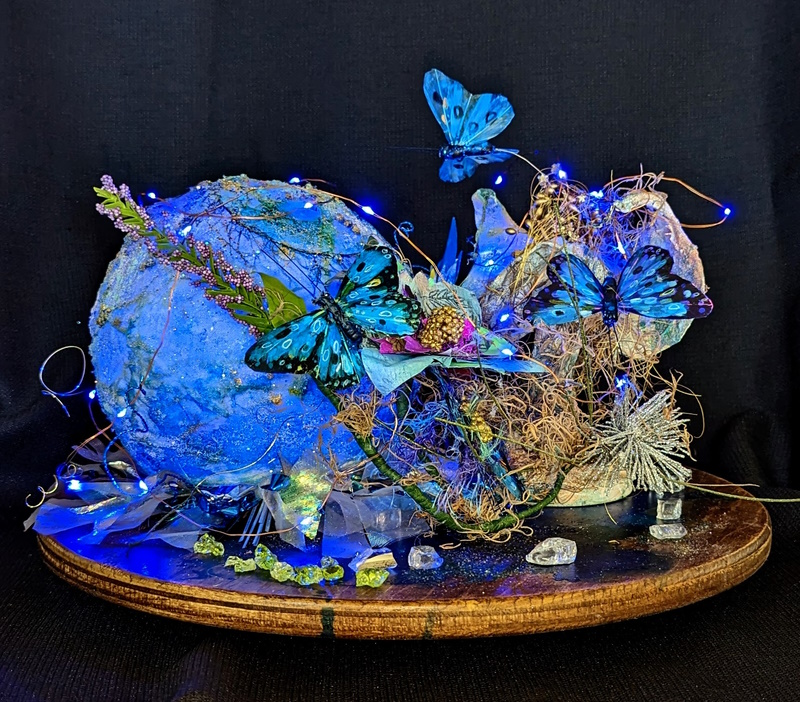 This is an image of a 3D sculpture on a custom made lazy susan. The sculpture is of an angel and floral elements around a LED lit orb made from cheesecloth and crochet fabric mixed with textile medium.