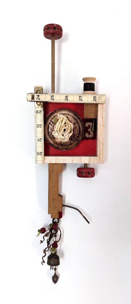 Assemblage sculpture of vintage sewing tools, a fabric flower, wooden pieces, beads, wire, and a small bell.