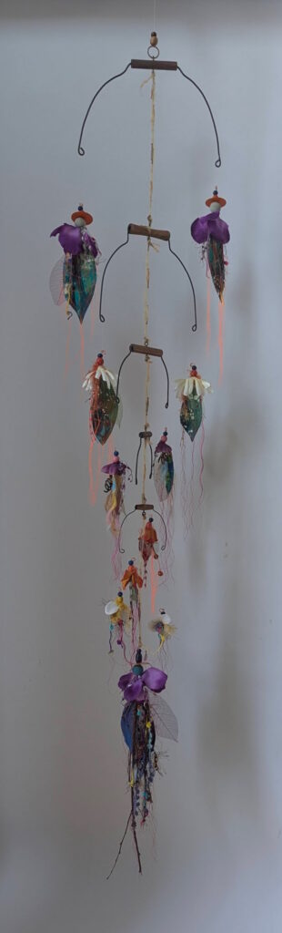 A suspended assemblage of curved steel forms, beads, translucent leaves, fabric, and trailing fibers cascades downward in a delicate arrangement of colorful, flower-like figures.