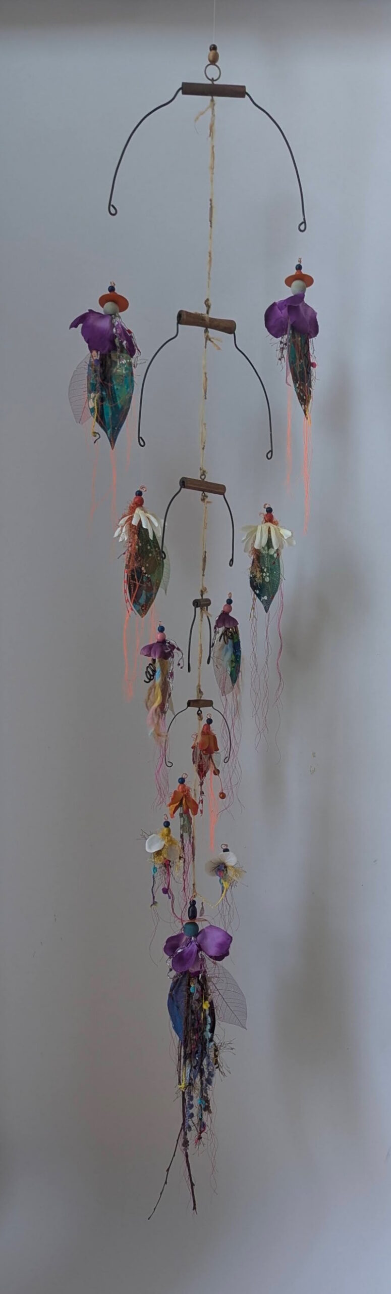 A suspended assemblage of curved steel forms, beads, translucent leaves, fabric, and trailing fibers cascades downward in a delicate arrangement of colorful, flower-like figures.